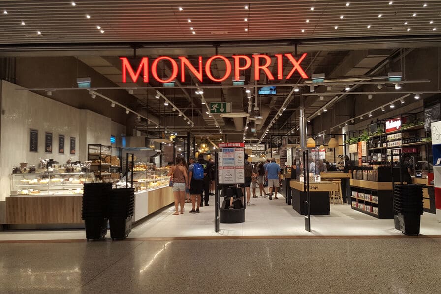 Entrée lumineuse d'un supermarché Monoprix situé dans une galerie commerciale, montrant les rayons frais, la zone de boulangerie et des clients qui circulent.