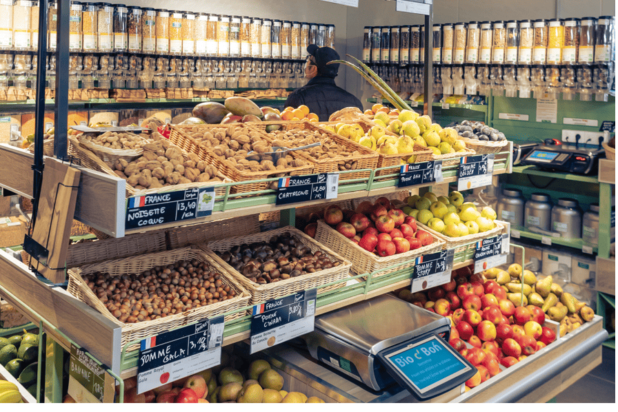 Étals de fruits et légumes frais en vrac dans un magasin Bio c' Bon, avec des pommes, des noisettes, des châtaignes et des distributeurs de produits secs en arrière-plan.