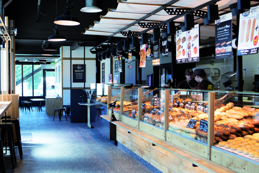 Vue intérieure d'une boulangerie Marie blachère montrant de larges vitrines remplies de pains, viennoiseries et pizzas, avec du personnel en arrière-plan.