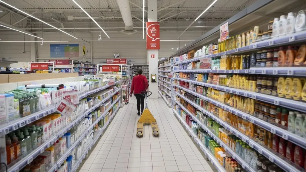 Vue en perspective d'une allée de supermarché Auchan. Une employée en uniforme rouge déplace un transpalette manuel jaune entre deux longs rayons remplis de produits.