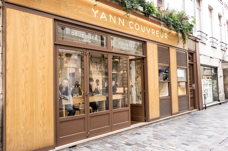 Façade en bois clair de la pâtisserie Yann Couvreur à Paris avec de grandes vitrines donnant sur une rue pavée.