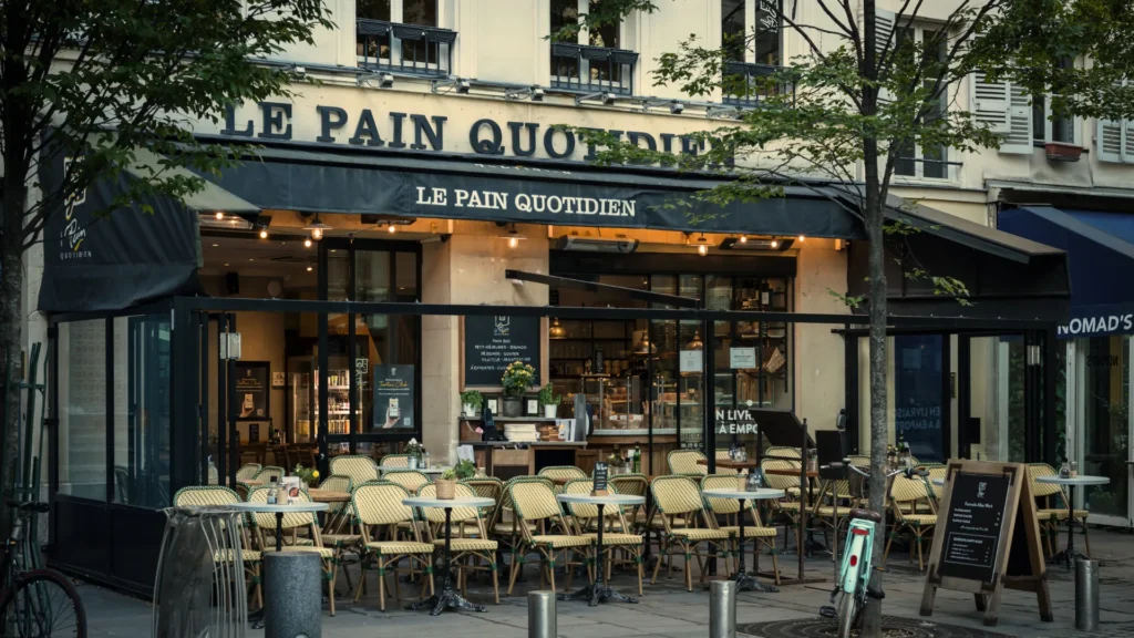 Terrasse extérieure du restaurant Le Pain Quotidien avec chaises en rotin et tables rondes sous un auvent noir.
