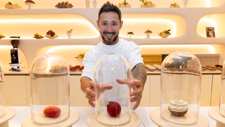 Le pâtissier Cédric Grolet souriant derrière un comptoir, présentant ses pâtisseries en trompe-l'œil (fraise, mangue, fleur) sous des cloches en verre dans une boutique au design épuré.