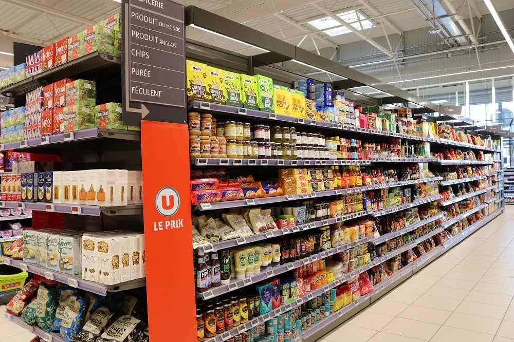 Allée d'un supermarché U montrant des étagères remplies de produits d'épicerie du monde, avec un panneau orange "U Le Prix" bien visible.
