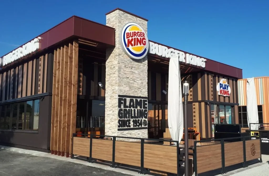 Façade moderne d'un restaurant Burger King avec une tour en pierre, une terrasse en bois et l'inscription 'Flame Grilling Since 1954' sous un ciel bleu clair.