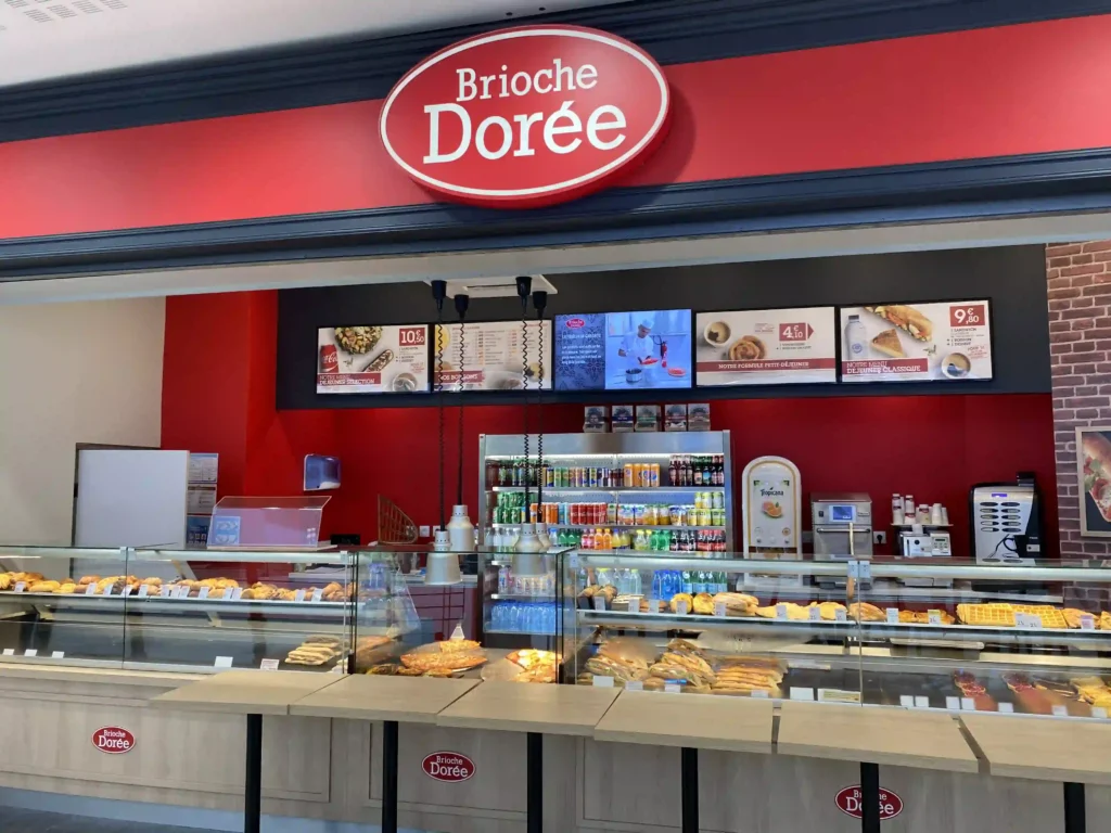 Comptoir de vente Brioche Dorée avec vitrines de sandwichs, pâtisseries et menus affichés sur écrans.
