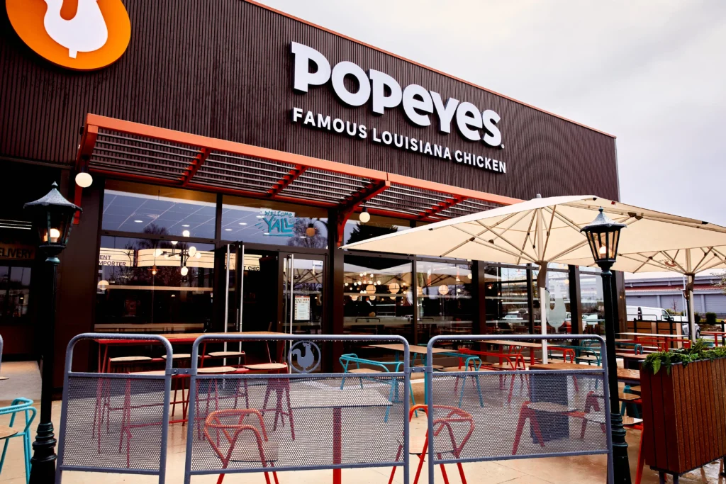 Façade extérieure d'un restaurant Popeyes Famous Louisiana Chicken avec une terrasse aménagée de parasols blancs et des chaises colorées.