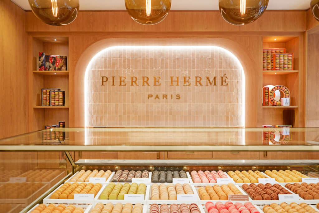 Présentoir de macarons colorés dans une boutique Pierre Hermé Paris au décor élégant en bois clair et carrelage mural beige.