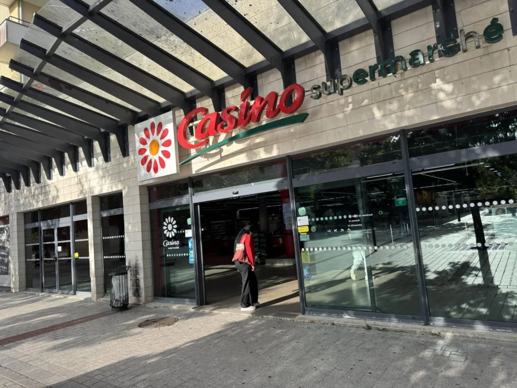 Entrée d'un supermarché Casino située sous un auvent moderne en verre et métal, avec une cliente s'apprêtant à entrer par les portes vitrées.