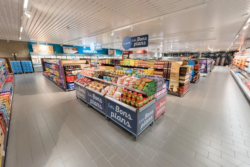 Intérieur d'un supermarché avec des présentoirs de promotion 'Les Bons Plans', rayons d'épicerie et surgelés, sol en carrelage gris