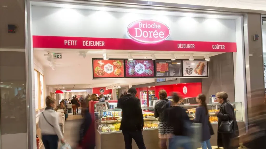 Entrée d'un restaurant Brioche Dorée avec clients devant le comptoir et panneaux affichant les offres Petit Déjeuner, Déjeuner et Goûter.