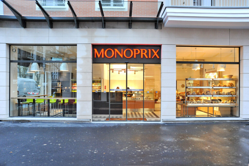 Façade moderne d'un magasin Monoprix avec de grandes baies vitrées laissant voir un espace boulangerie et un coin restauration avec des chaises hautes vertes.