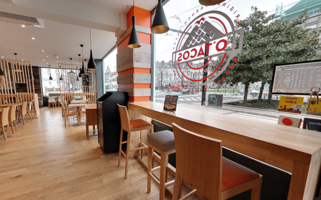 Intérieur design et chaleureux du restaurant O'Tacos avec mobilier en bois, comptoir face à la fenêtre et décoration aux couleurs orange et grise.
