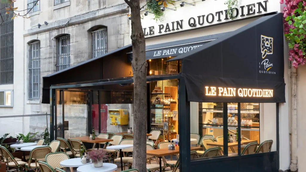 Façade d'un établissement Le Pain Quotidien rue des Archives avec terrasse abritée et éclairage chaleureux.