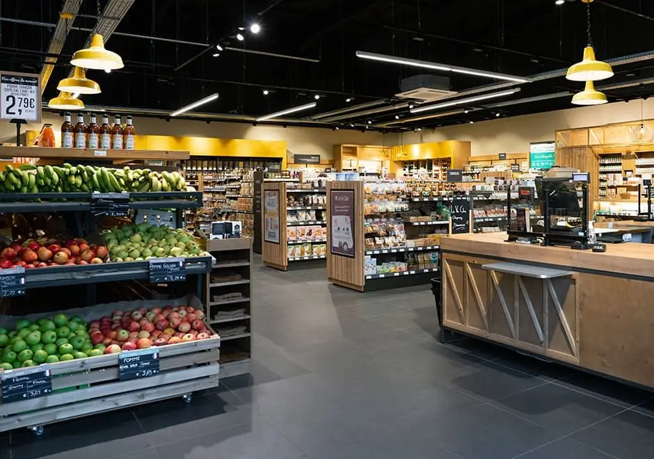 Rayon fruits et légumes à l'entrée d'un magasin La Vie Claire avec des pommes, bananes et poires bien rangées sous des luminaires jaunes.