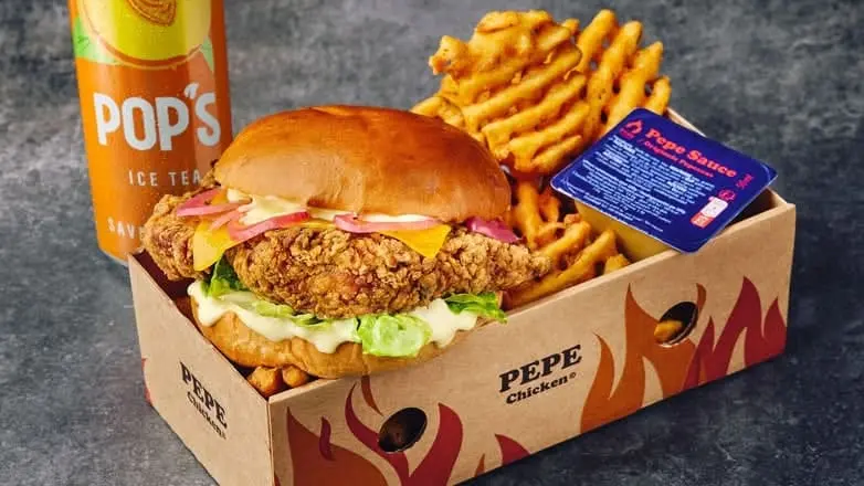 Un burger au poulet frit croustillant de chez Pepe Chicken avec oignons rouges et fromage, servi avec des frites gaufrées, une sauce Pepe Sauce et une canette de thé glacé.