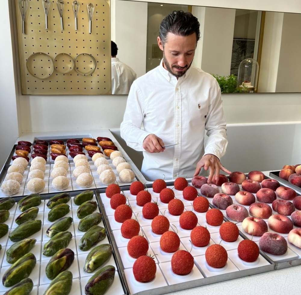 Cédric Grolet en chemise blanche préparant des plateaux de pâtisseries trompe-l'œil en forme de fruits : pêches, fraises, citrons et pistaches alignés avec précision.