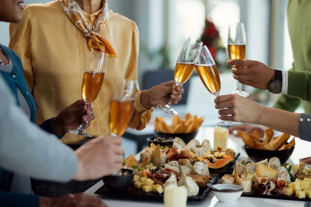 Un groupe de collègues porte un toast avec des verres de cidre et de vin pétillant lors d'une fête d'entreprise. Si vous vous demandez quel alcool peut-on boire sans gluten, ces options sont d'excellents choix pour accompagner un buffet de fromages et de charcuteries.