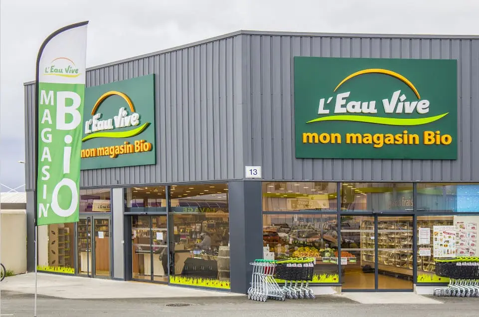Extérieur du magasin bio L'Eau Vive avec enseigne 'mon magasin Bio', drapeaux publicitaires et caddies à l'entrée.