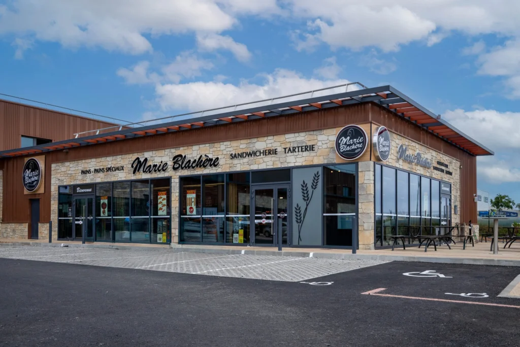 Façade moderne d'une boulangerie Marie Blachère avec son logo, une terrasse extérieure et un parking sous un ciel bleu.