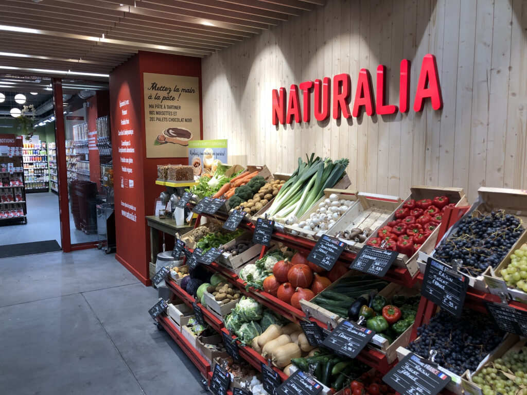 Intérieur d'un magasin Naturalia présentant un étalage de légumes frais dans des cagettes en bois : poireaux, poivrons rouges, courges et raisins, sous une enseigne rouge sur mur en bois.