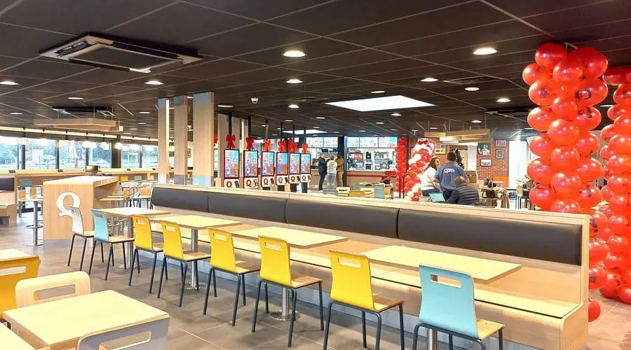 Vue intérieure d'un restaurant Quick moderne avec des rangées de tables en bois, des chaises jaunes et bleues, une longue banquette grise et des bornes de commande tactiles en arrière-plan.