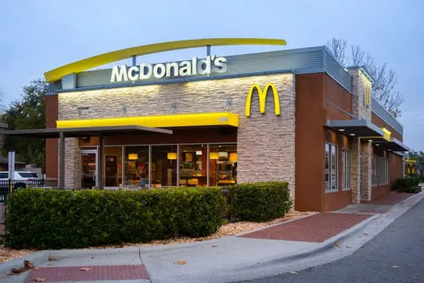 Façade extérieure d'un restaurant McDonald's moderne au crépuscule, présentant des murs en pierre apparente, le logo emblématique de l'arche dorée illuminé et une haie verdoyante au premier plan.