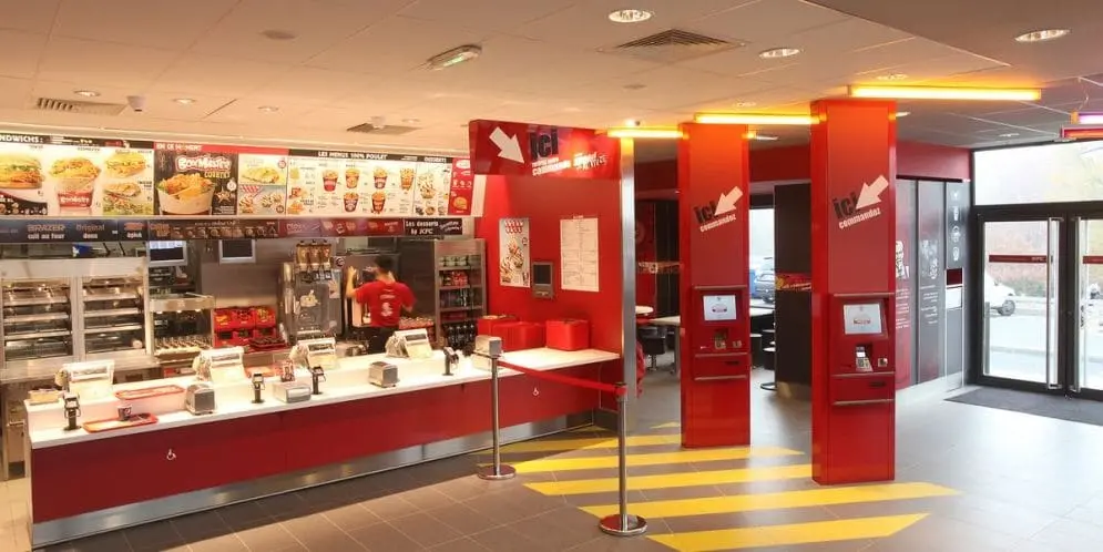 Intérieur d'un restaurant KFC avec bornes de commande automatiques et comptoir de service.