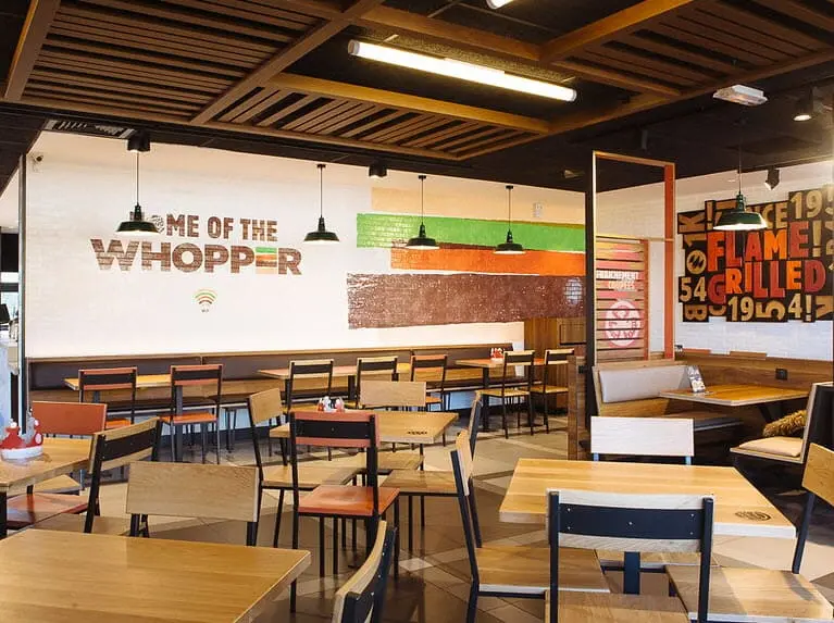 Intérieur d'une salle de restaurant Burger King avec du mobilier en bois, des luminaires suspendus noirs et un mur décoré du slogan 'Home of the Whopper'