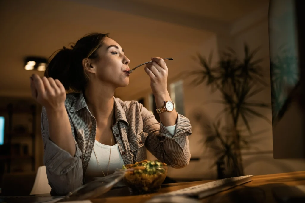 Une femme de profil savoure une salade à son bureau le soir, les yeux fermés, devant un écran d'ordinateur allumé.