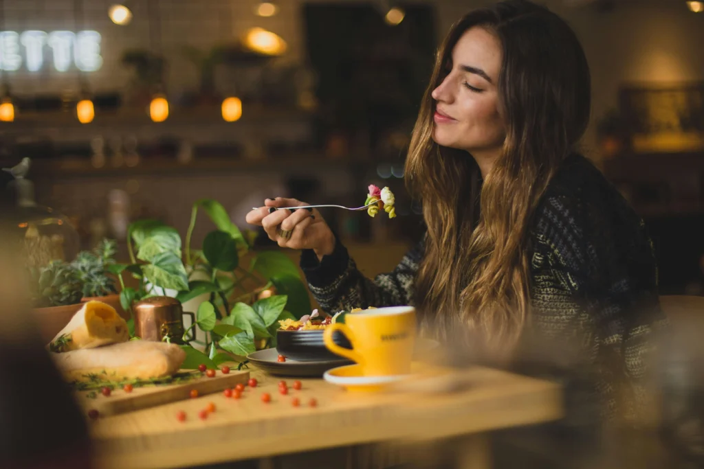 Une femme aux longs cheveux châtains savoure une bouchée de pâtes colorées, les yeux fermés, attablée dans un restaurant à l'ambiance chaleureuse et tamisée avec des plantes vertes en arrière-plan.