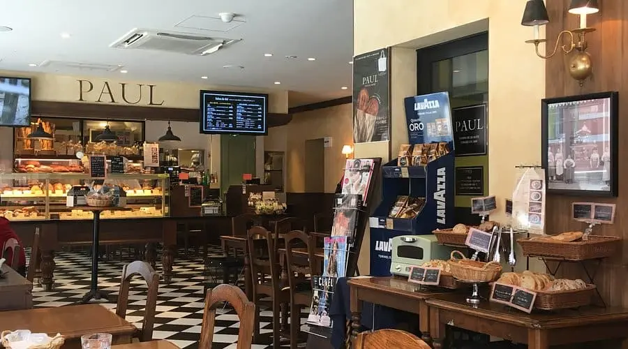 Intérieur d'une boulangerie Paul avec sol à damier noir et blanc, tables en bois, présentoirs à pain et comptoir de vente.