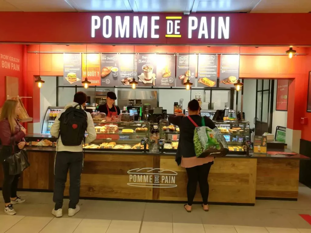 Comptoir de vente Pomme de Pain à Lille avec clients en attente et affichage des offres de sandwichs et burgers.