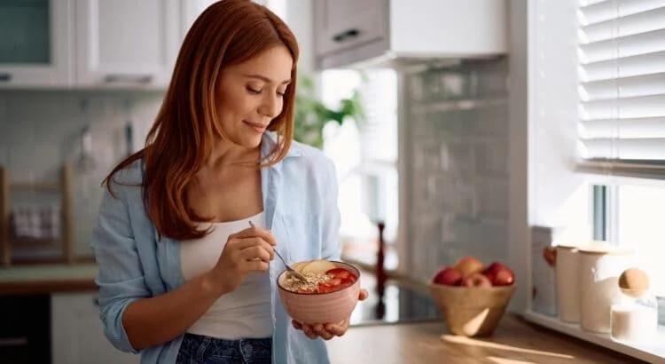 Femme rousse souriante dans une cuisine lumineuse, tenant un bol de porridge aux céréales garni de tranches de pommes et de fraises.