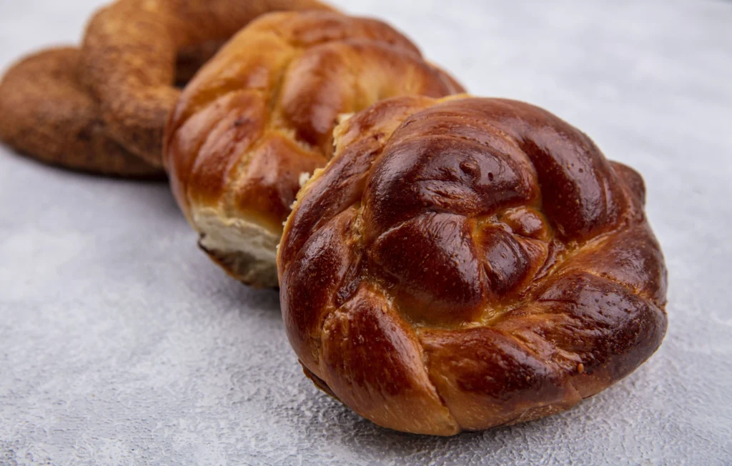 Deux brioches rondes dorées et tressées au premier plan avec des simits turcs en arrière-plan sur une surface grise.