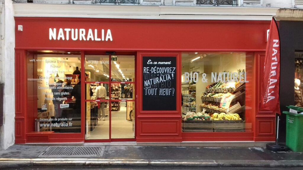 Façade rouge vif d'un magasin bio Naturalia avec de grandes vitrines laissant voir des étals de fruits et légumes à l'intérieur.