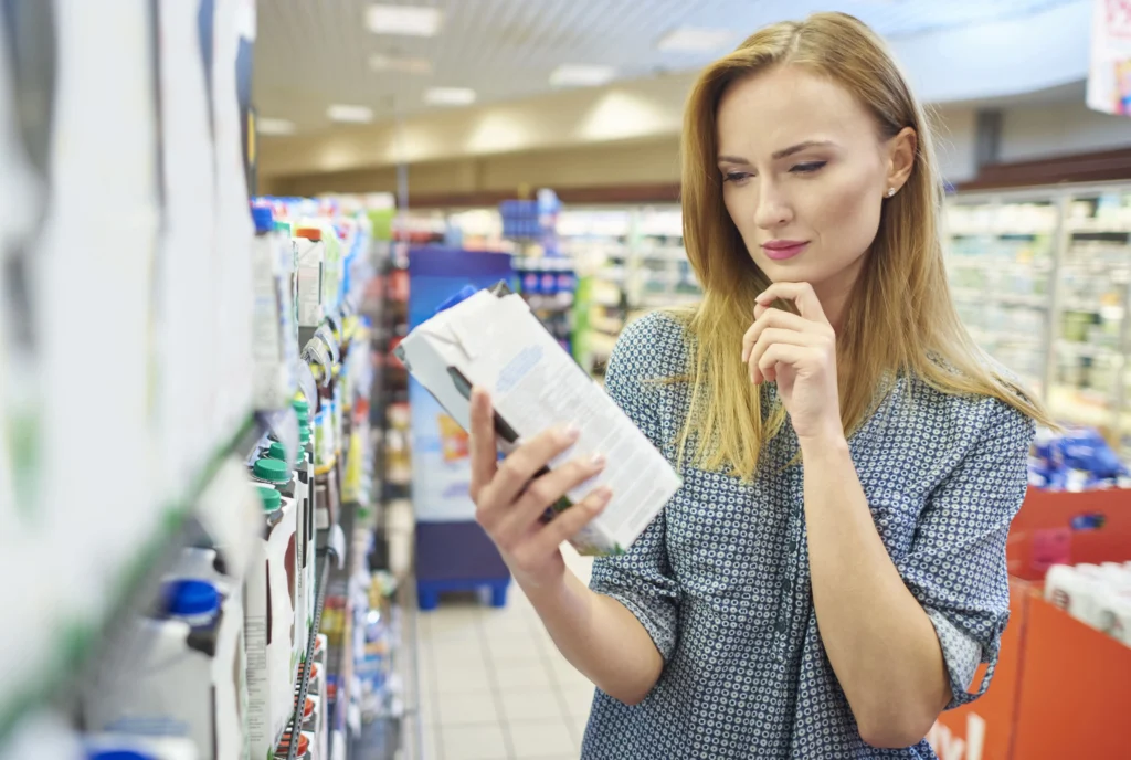 Femme blonde pensive lisant les ingrédients sur une brique de boisson dans le rayon frais d'un supermarché.