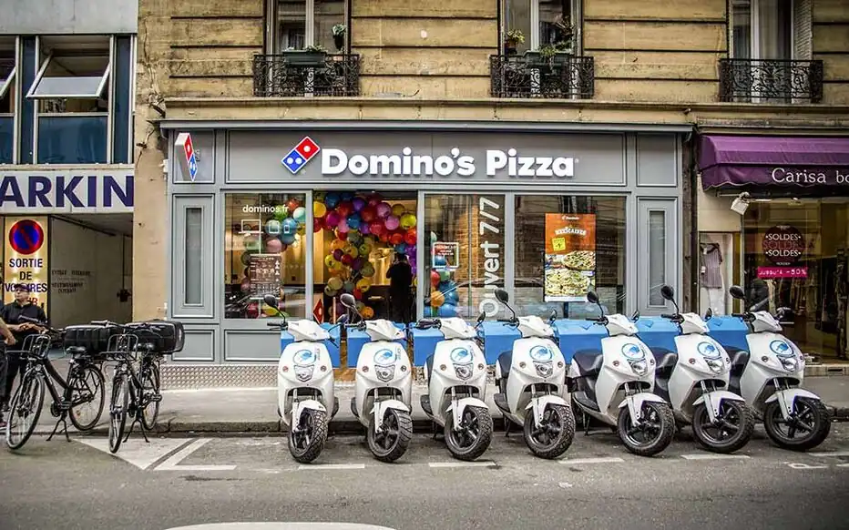 Devanture d'un restaurant Domino's Pizza en ville avec flotte de scooters de livraison garés devant